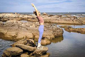 Woman stands on rocky coast holding a yoga pose with arms raised, a Health & Medical Photo by DanFLCreativo