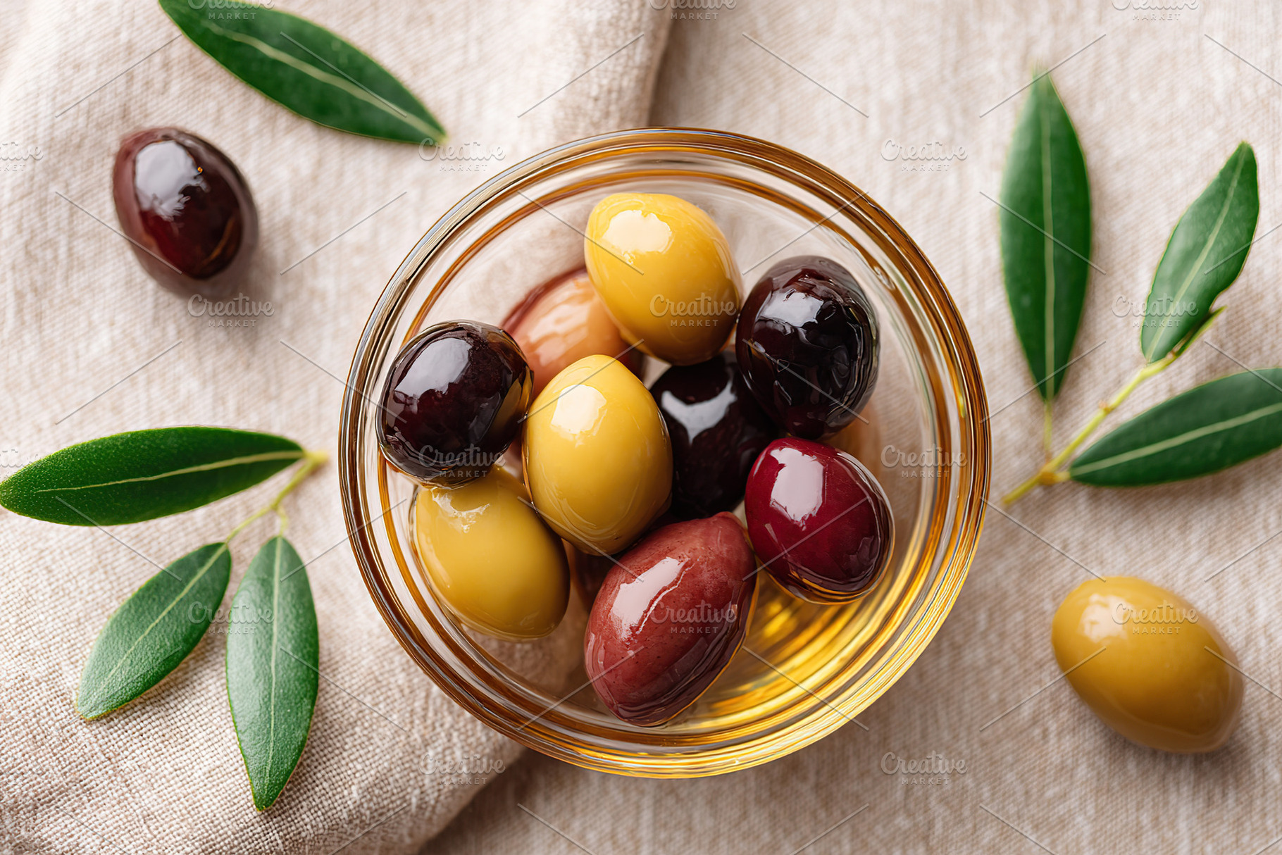 Assorted marinated olives arranged in a glass bowl with olive leaves on a cloth, a Food & Drink Photo by AlyaAnd