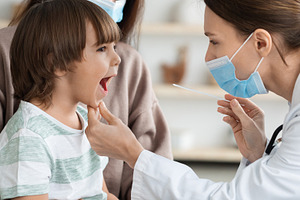 Professional woman pediatrician wearing protective face mask checking throat of, a Photo by Prostock-Studio