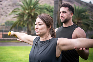 Male personal trainer assisting woman with dumbbell exercise outdoors