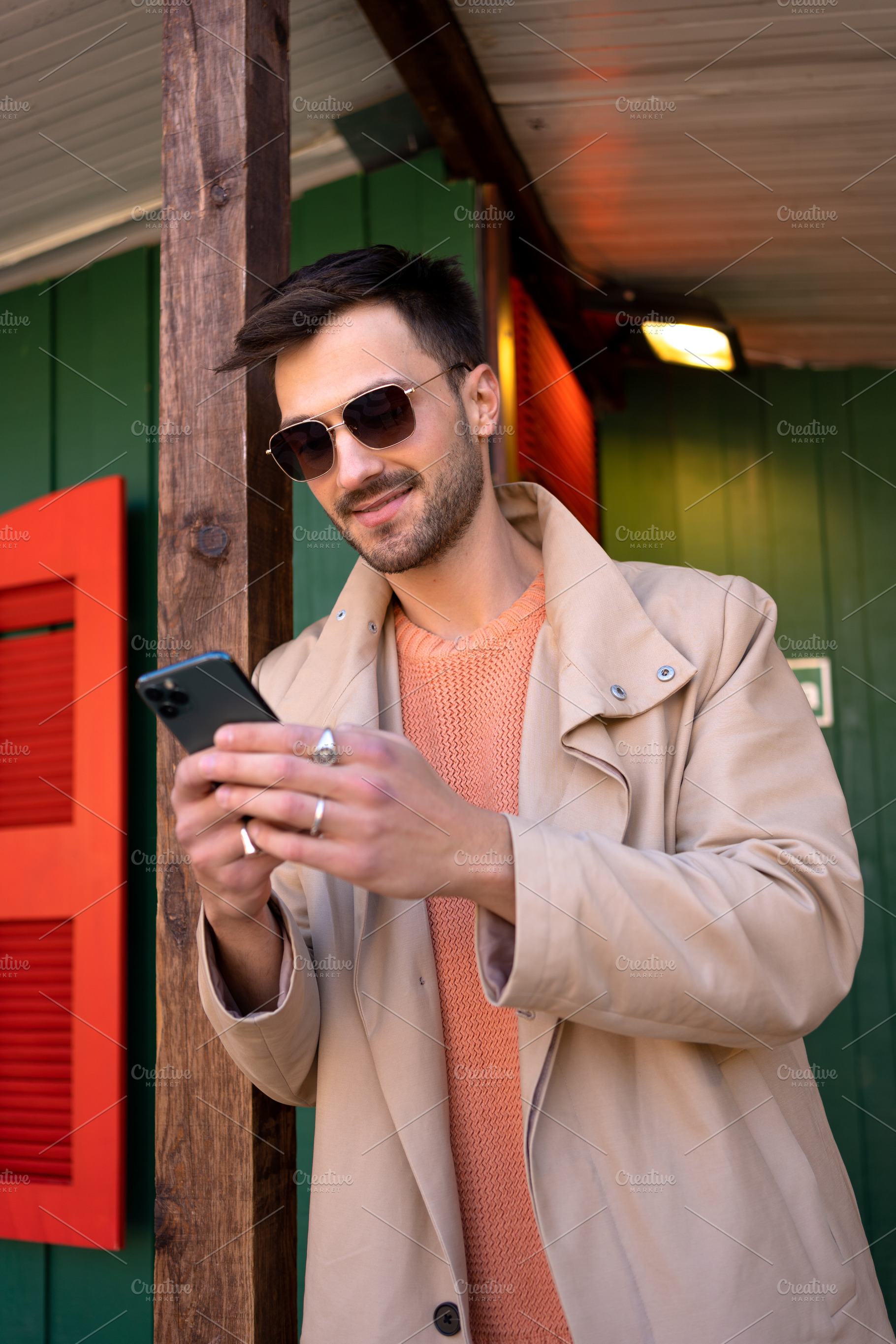 Smiling man in beige trench coat using smartphone outside green house, a Person Photo by andreonegin market