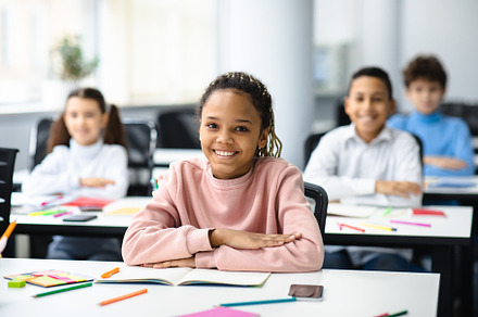 Portrait of small black girl sitting at desk in classroom, a Photo by Prostock-Studio