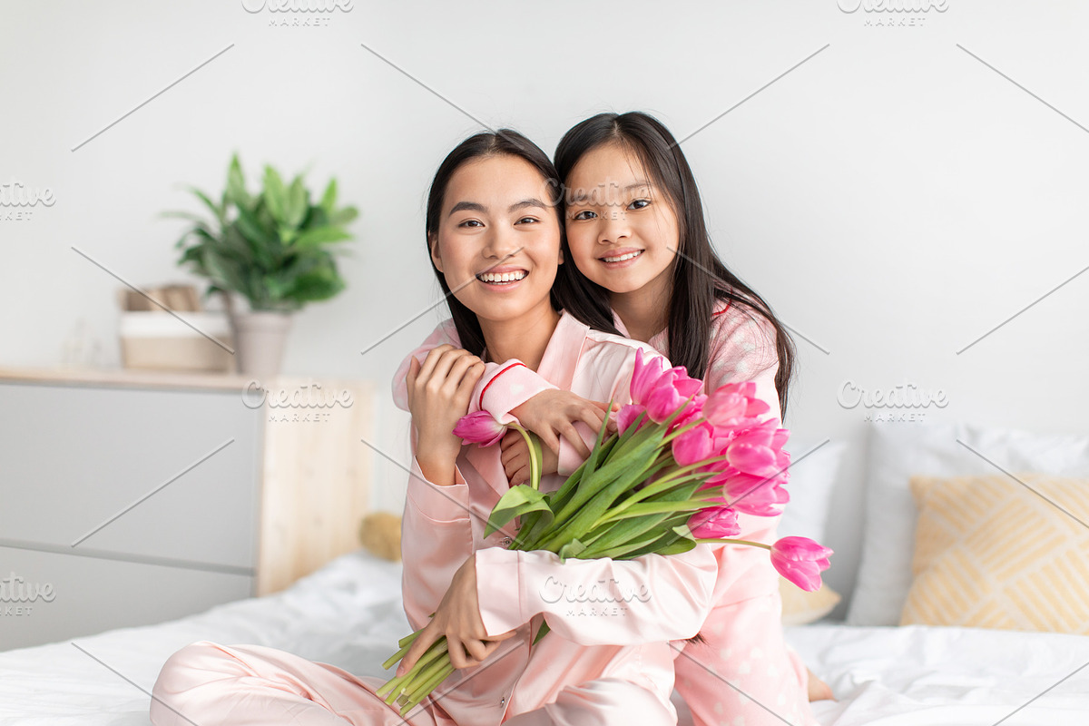 Cheerful chinese young girl hug millennial lady in pink pajama with tulips bouq, a Photo by Prostock-Studio (Photo 1 of 2)