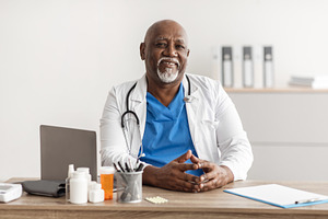 Portrait of smiling doctor looking at camera sitting at desk, a Photo by Prostock-Studio