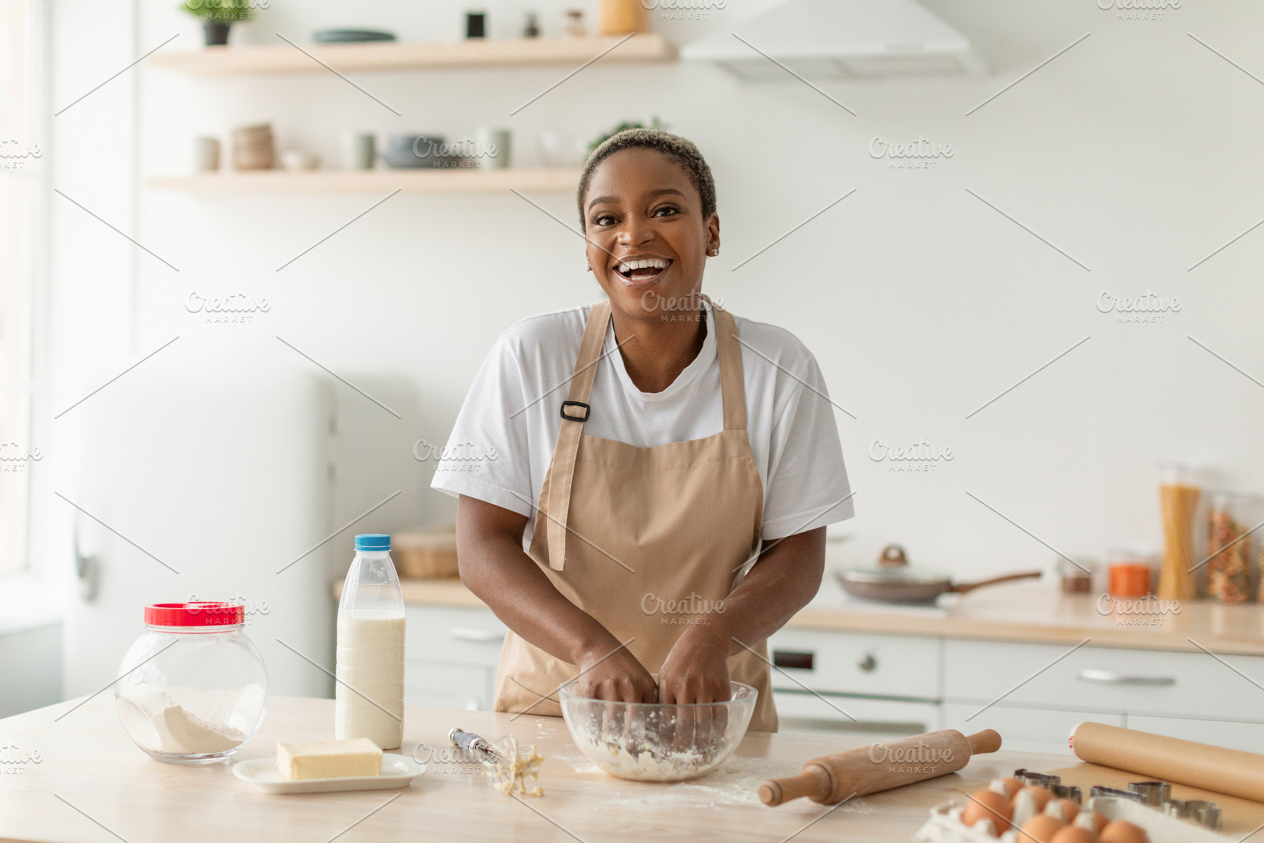 Laughing young african american lady in apron making pie dough in minimalist ki, a Photo by Prostock-Studio