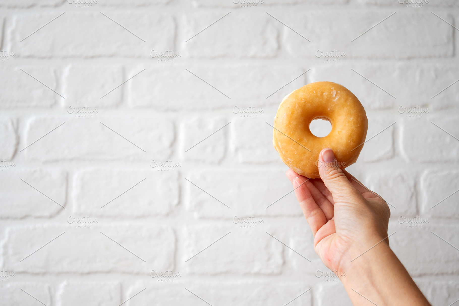 Hand Holding Glazed Donut with Copy Space on White Brick Background, a Food & Drink Photo by etorres69