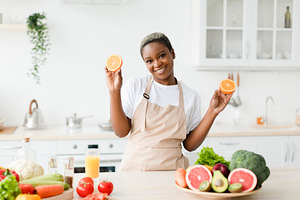 Satisfied young pretty black female in apron cooking, shows fresh oranges in ki, a Person Photo by Prostock-Studio