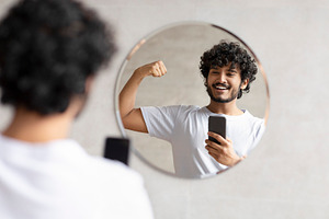 Excited indian man taking photo on smartphone in bathroom mirror, showing perfe, a Person Photo by Prostock-Studio