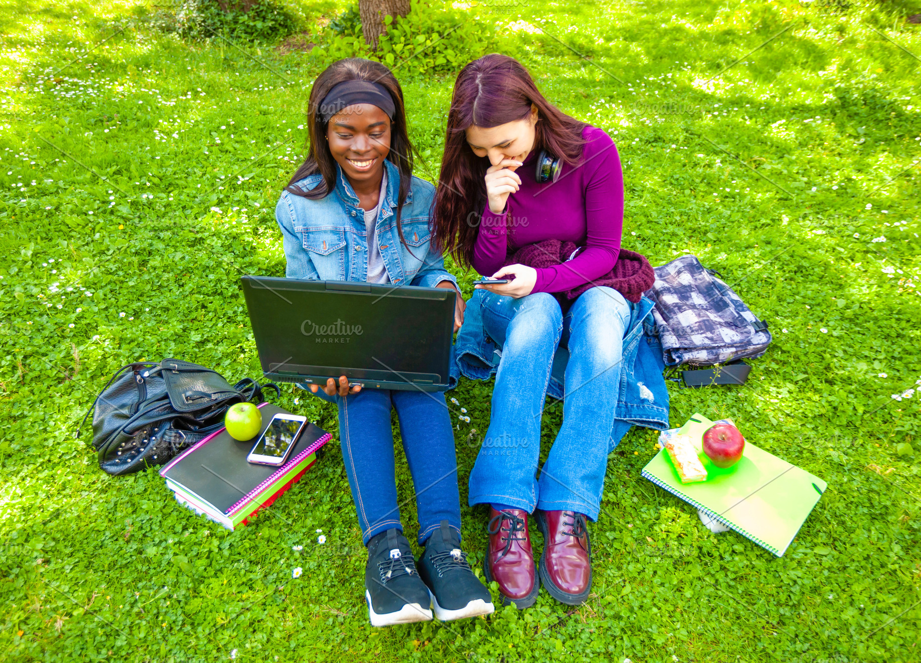Two students using laptop and smartphone sitting on grass in a park, a Person Photo by Antonio Gravante Market