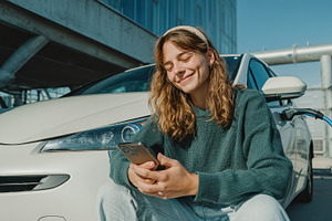 Woman using phone near charging elec, a Photo by Pixeness Studio