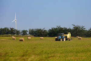 Modern tractor baling hay on a green field with round bales and a wind turbine, an Industrial Photo by Ales Munt