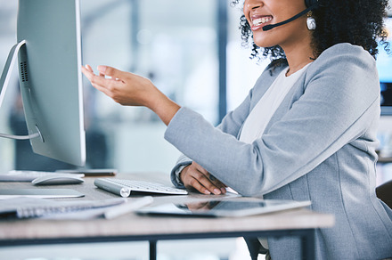 Closeup of one happy young mixed race call centre telemarketing agent talking on a headset while working on a computer in an office. Confident friendly female consultant operating helpdesk for customer service and sales support, a Photo by Peopleimages.com