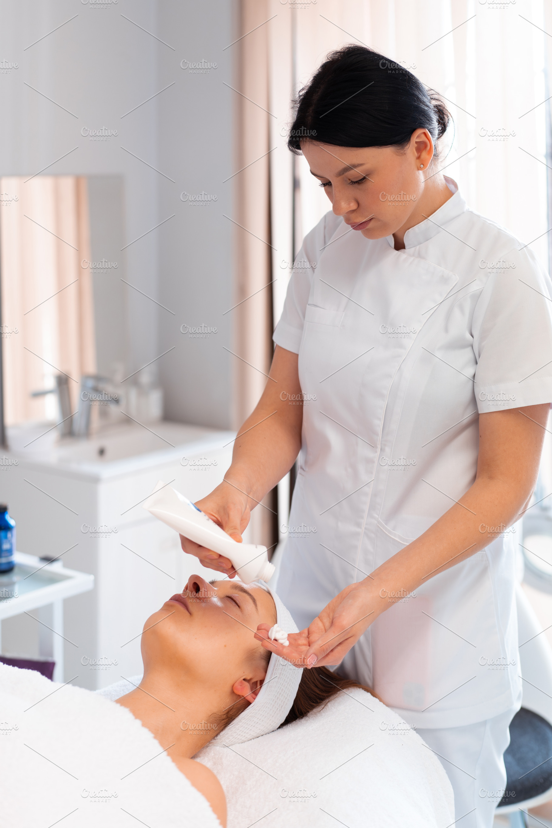 Masseuse taking cream in hand from bottle while massaging woman's face, a Person Photo by andreonegin market