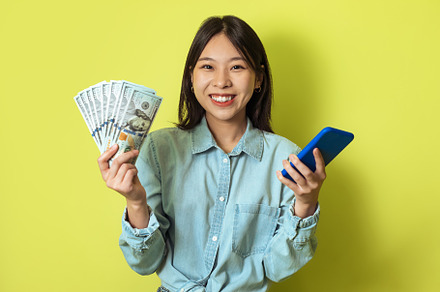 Cheerful Asian Lady Holding Smartphone And Money Cash, Yellow Background, a Person Photo by Prostock-Studio