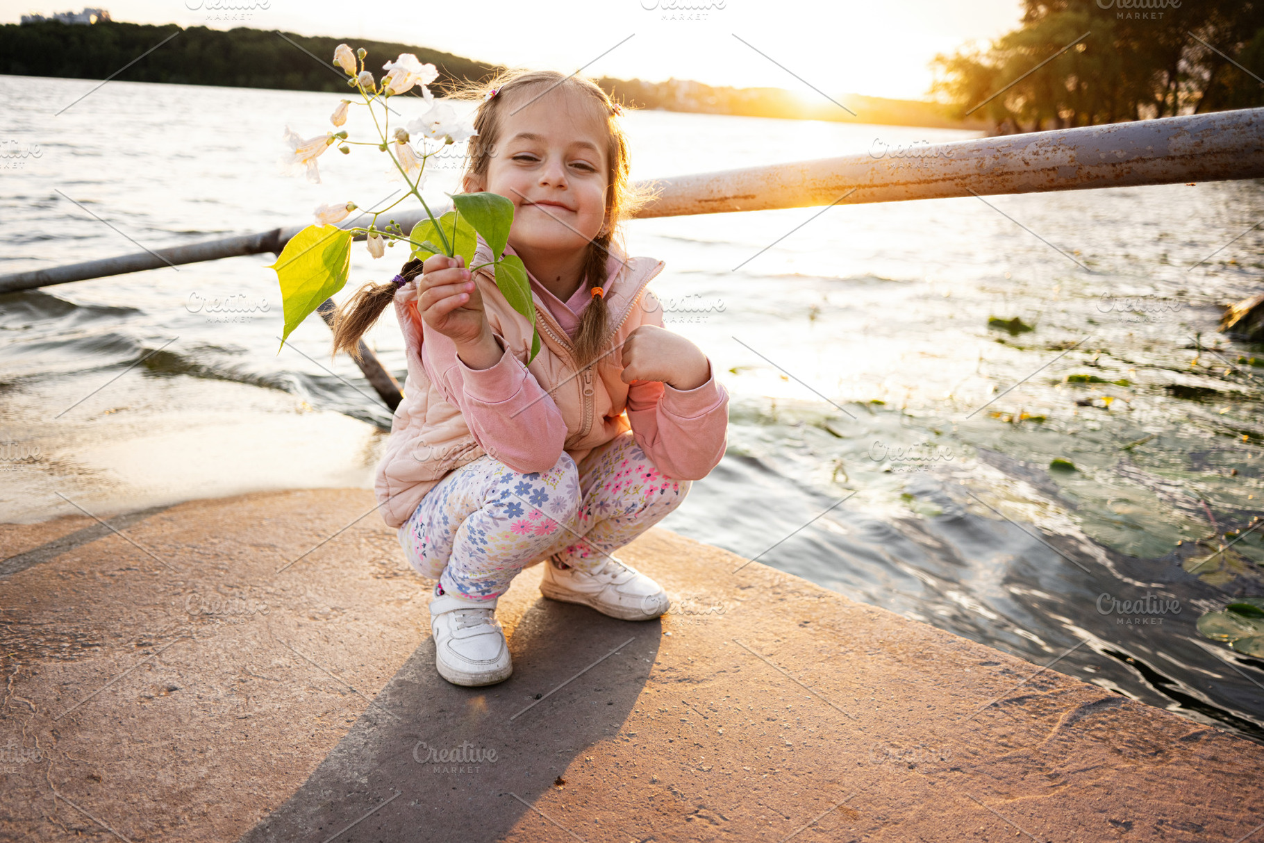 Young girl enjoying sunset by a lake holding flowers with a joyful smile, a Photo by AS photostudio
