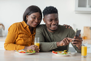 Happy African American Couple Relaxing With Smartphone While Having Breakfast I, a Photo by Prostock-Studio