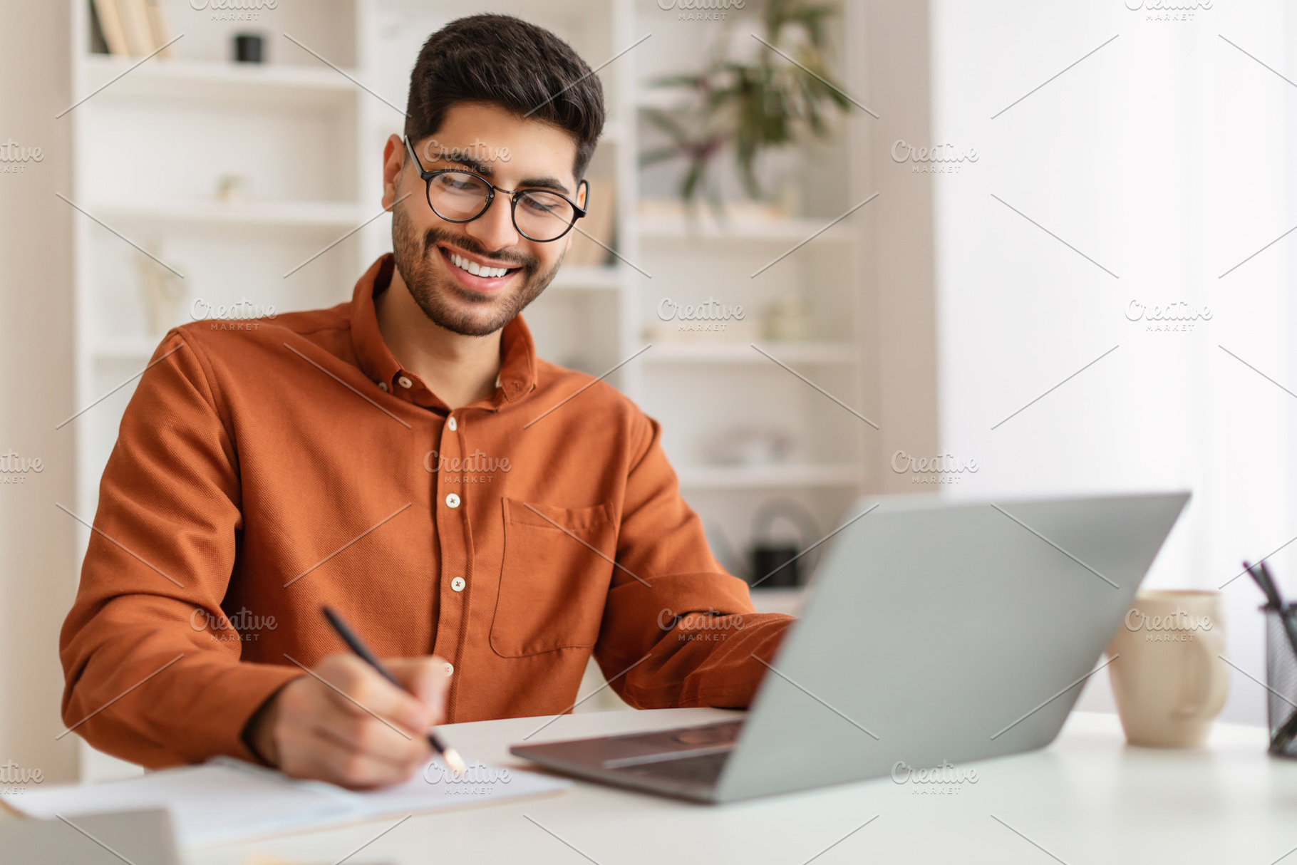 Smiling Arab man in glasses using laptop and taking notes, a Photo by Prostock-Studio