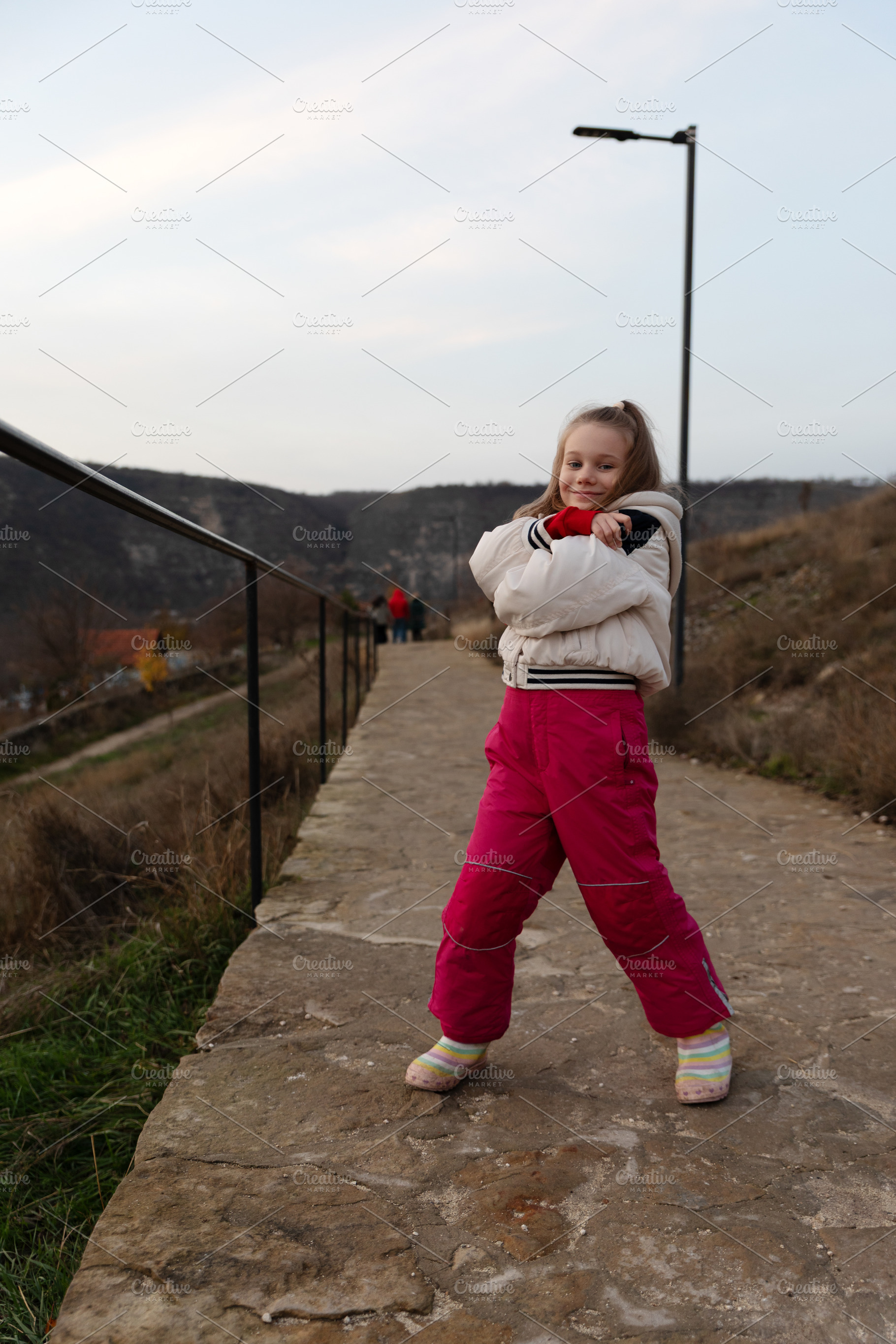 Cheerful young girl in a white jacket and pink pants standing on, a Person Photo by Ales Munt
