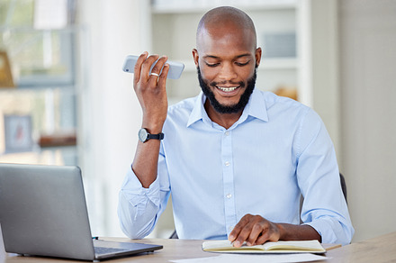 Young african american businessman on a call using a phone while reading a note in a notebook and working on a laptop in an office at work alone. One male business professional talking on a cellphone while working, a Photo by Peopleimages.com