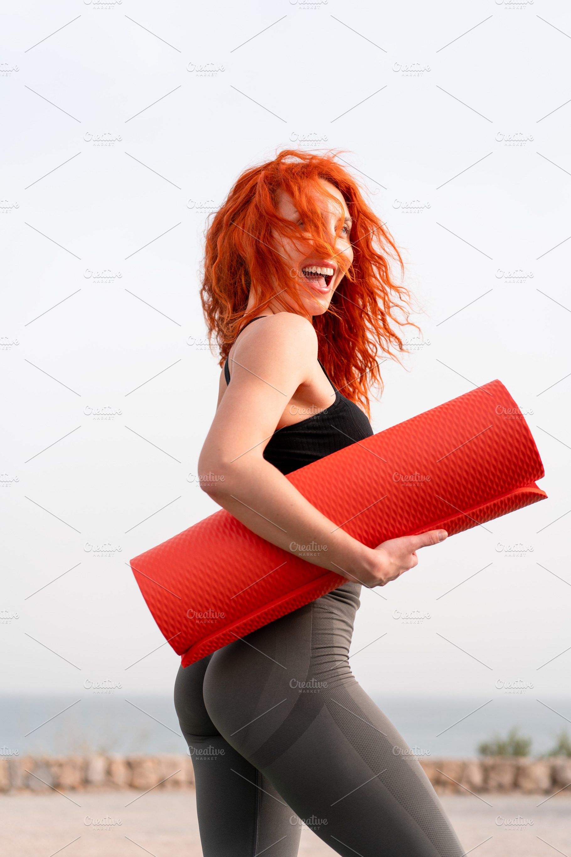 Cheerful sporty woman holding rolled up exercise mat against clear sky, a Photo by andreonegin market