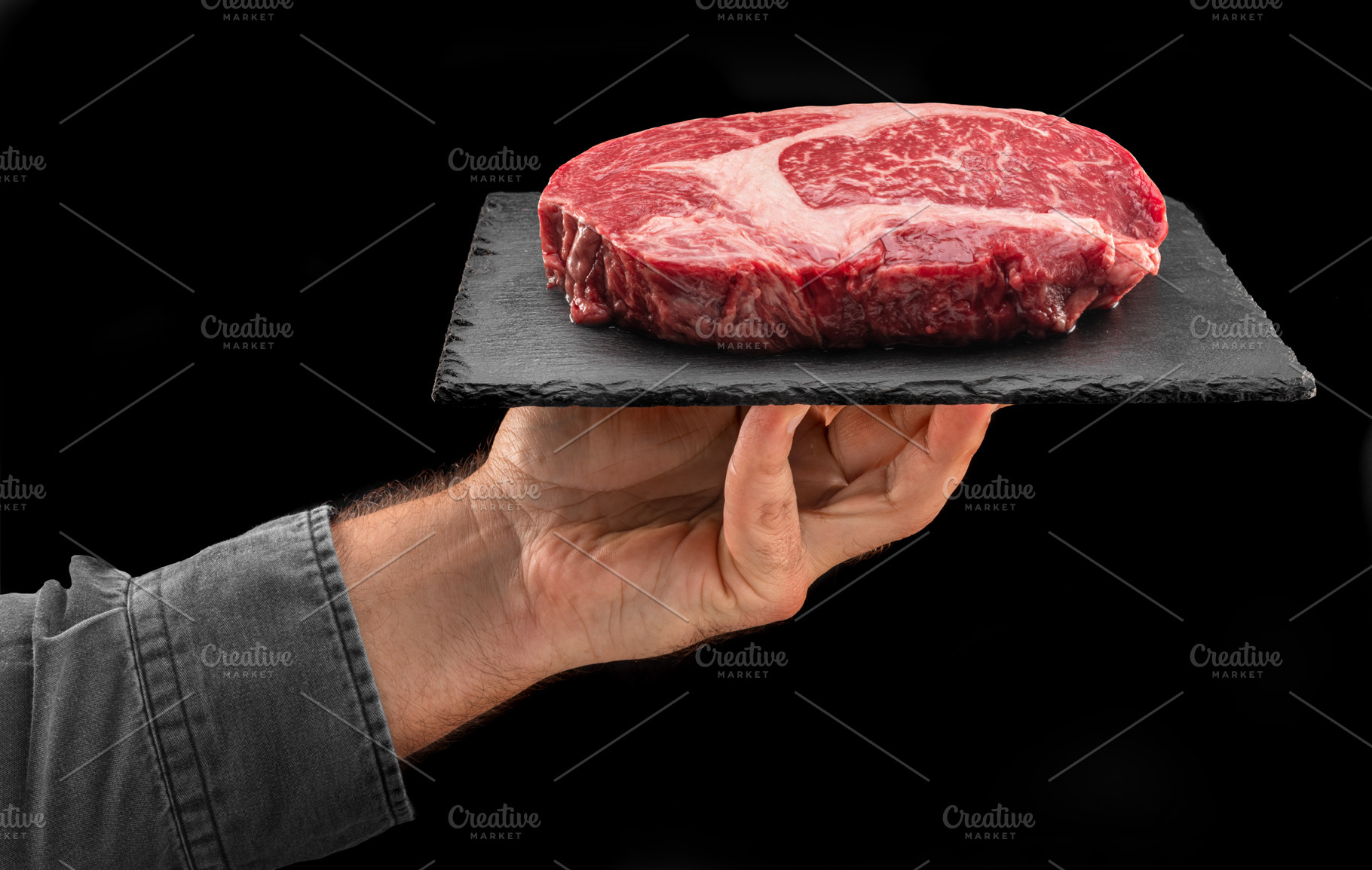 Cook holds graphite serving board with raw ribeye steak on black, a Food & Drink Photo by Volff