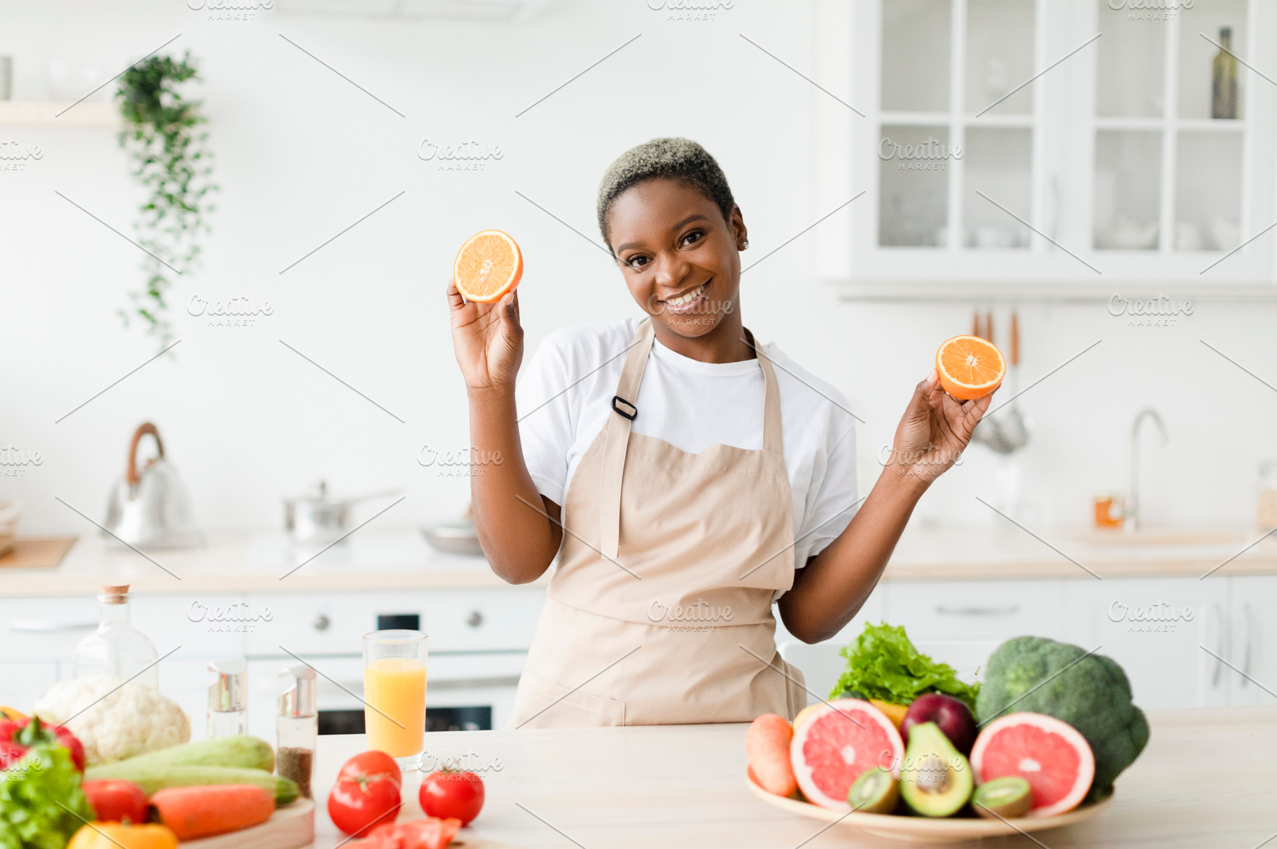 Satisfied young pretty black female in apron cooking, shows fresh oranges in ki, a Person Photo by Prostock-Studio