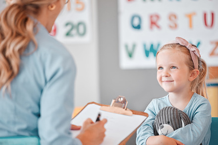 Young smiling caucasian child sitting with her psychologist in a clinic. Cute girl talking to a mental health professional. Using a clipboard to take notes on her patient. Dyslexia and anxiety, a Photo by Peopleimages.com