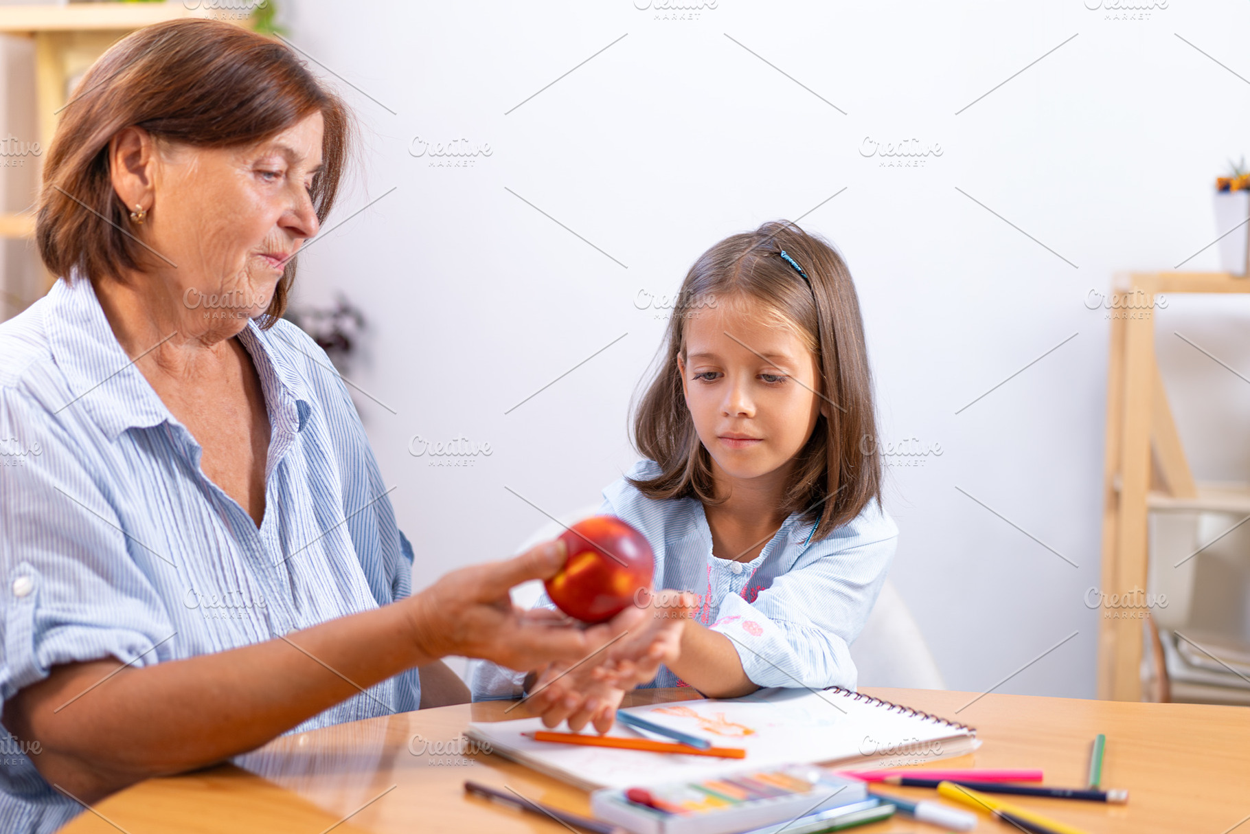 Grandmother placing apple on girl's hand over colored pencils and book, a Photo by andreonegin market