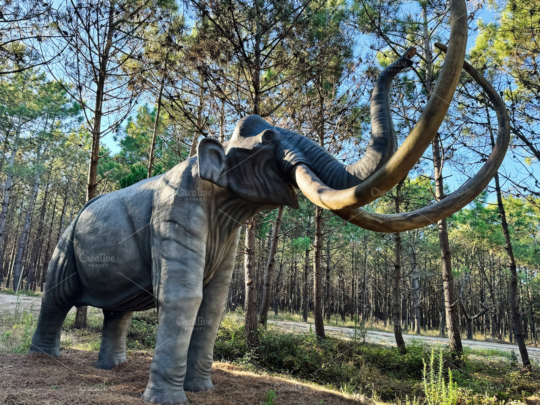Life size mammoth sculpture amongst trees in a lush forest setting, a Photo by AS photostudio