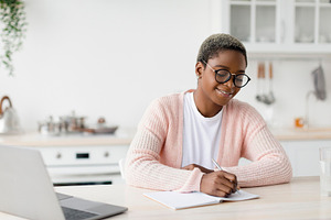 Cheerful millennial pretty black female student or worker in glasses at table w, a Person Photo by Prostock-Studio
