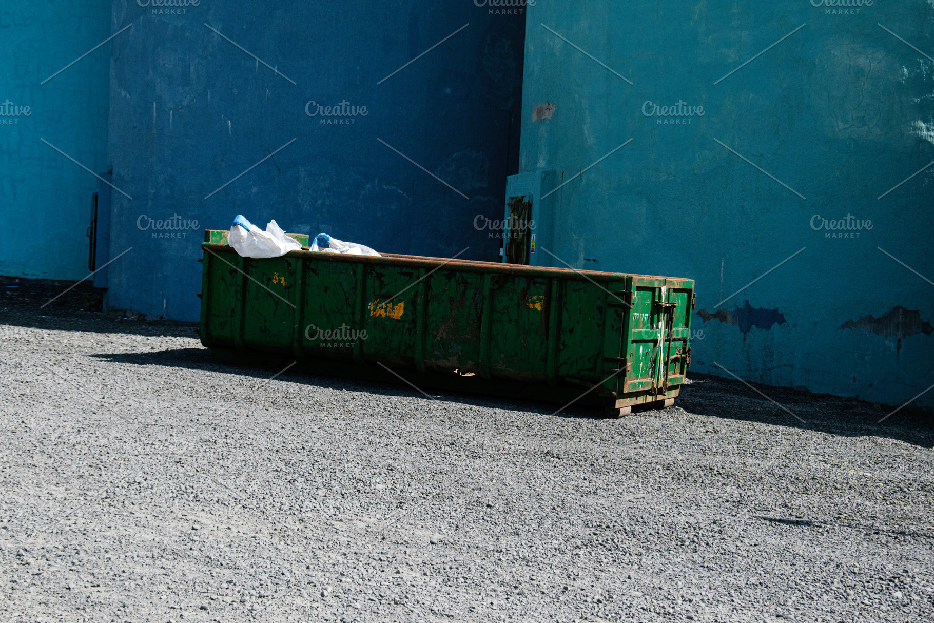 Green dumpster against blue walls in a sunlit area in Akranes, Iceland, a Photo by AS photostudio