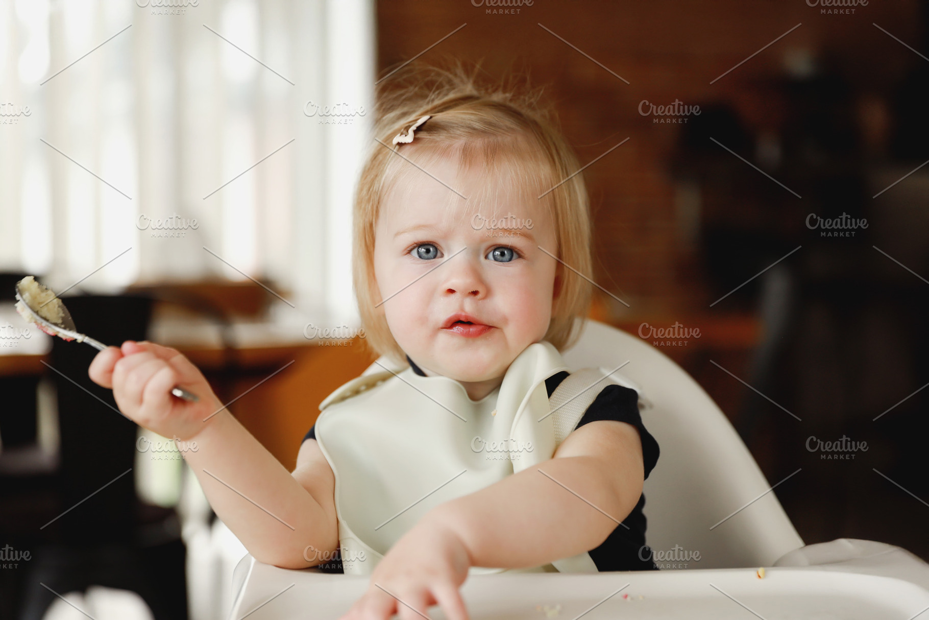 Infant girl or toddler sitting on high chair. Eating food by herself using spoo, a Person Photo by Lenor4ik