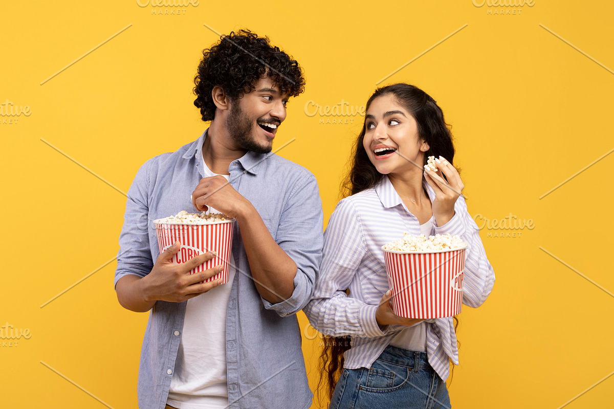 Overjoyed indian man and woman eating popcorn, standing back to back and smilin, a Photo by Prostock-Studio (Photo 2 of 5)