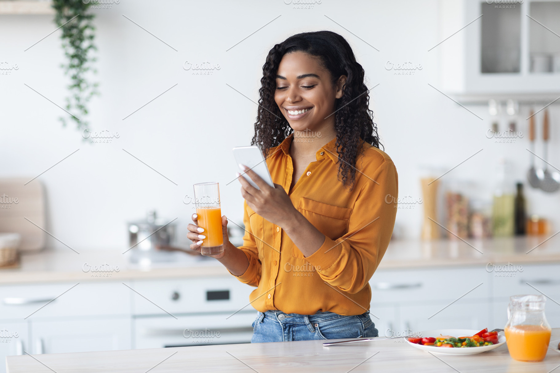 Positive pretty black lady holding glass of juice and smartphone, a Photo by Prostock-Studio