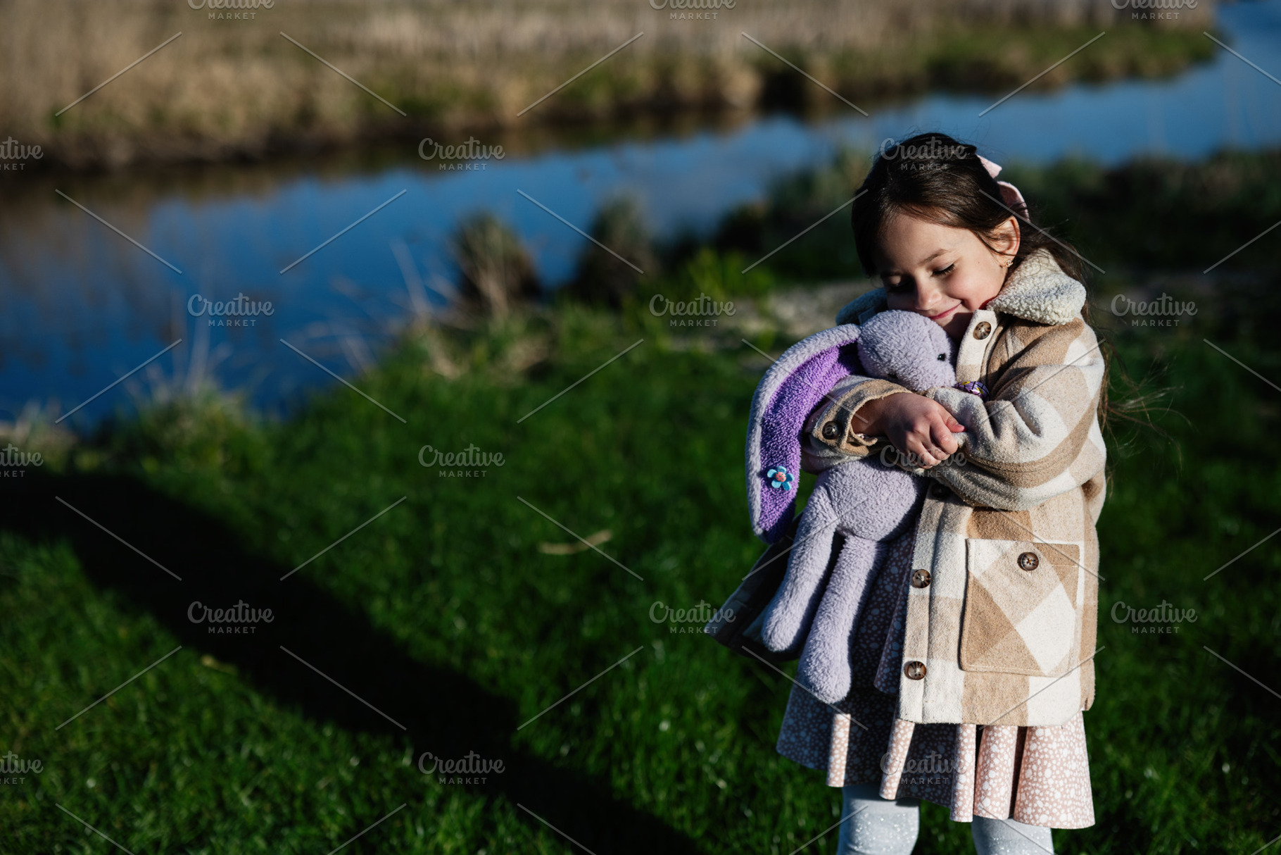 Little girl hugging a purple plush bunny outdoors on a grassy riverbank wearing, a Person Photo by AS photostudio