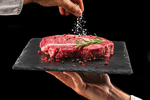 Chef is salting or seasoning raw ribeye steak laying on graphite, a Food & Drink Photo by Volff