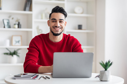 Portrait Of Smiling Handsome Arab Male Freelancer Posing At Desk With Laptop, a Photo by Prostock-Studio