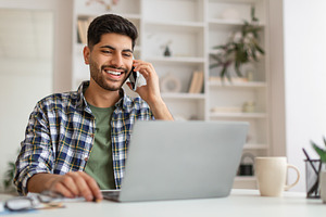 Smiling Arab man working and talking on phone at home, a Photo by Prostock-Studio