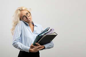 Stressed female office worker with heap of papers talking on smartphone on ligh, a Photo by Prostock-Studio