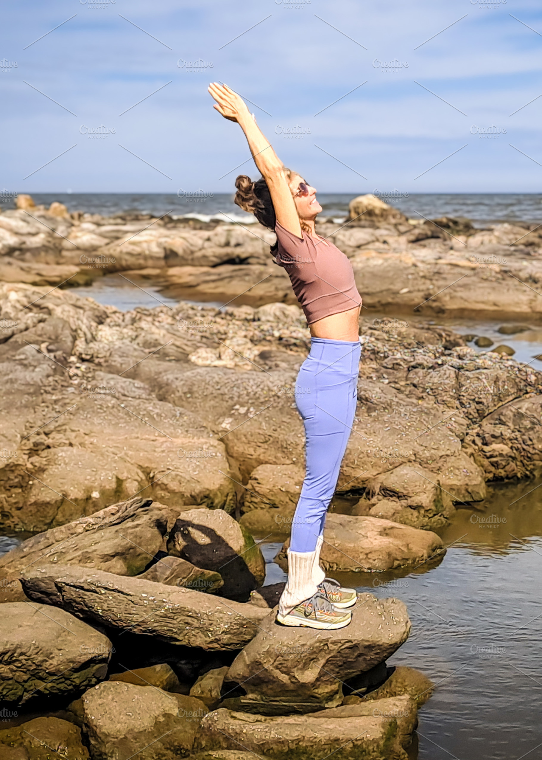Woman stands on rocky coast holding a yoga pose with arms raised, a Health & Medical Photo by DanFLCreativo