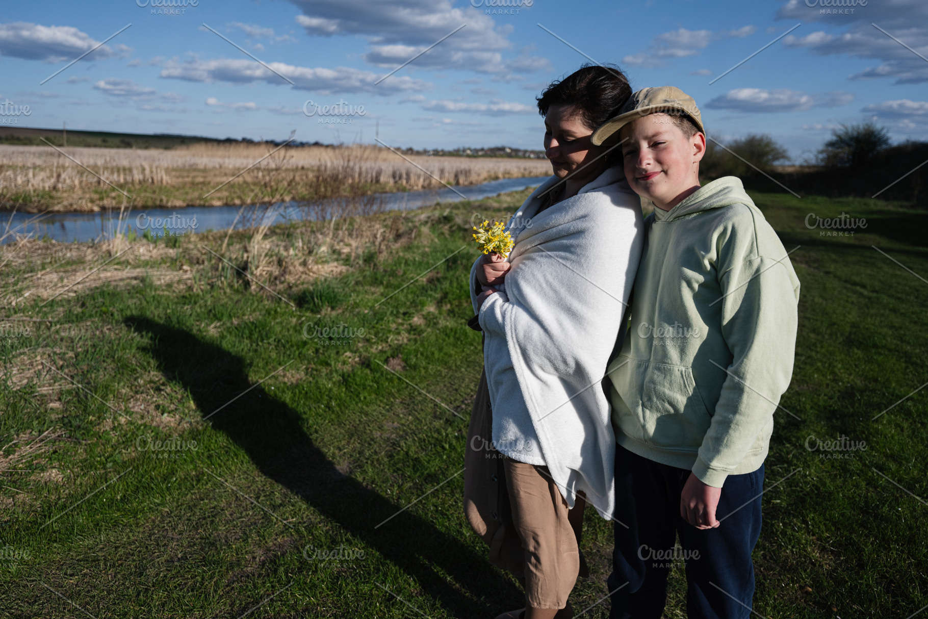 Mother and son wrapped in a blanket by the riverside holding yellow wildflowers, a Person Photo by AS photostudio