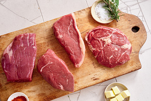 Raw meat cuts displayed on wooden board with seasoning and herbs at kitchen cou