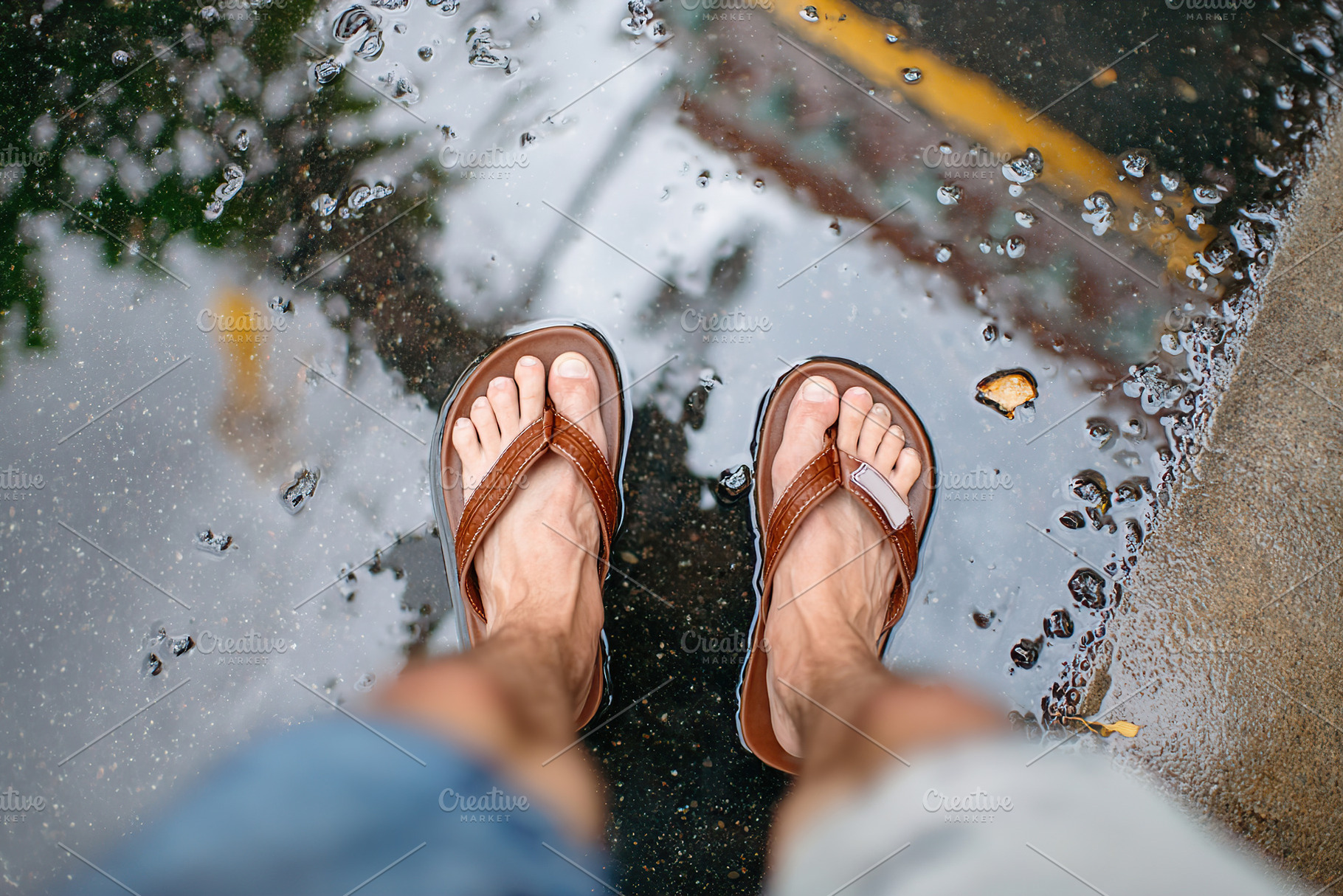 Wet pavement reflects sandals worn by a person in casual attire on a rainy day, an Abstract Photo by AlyaAnd