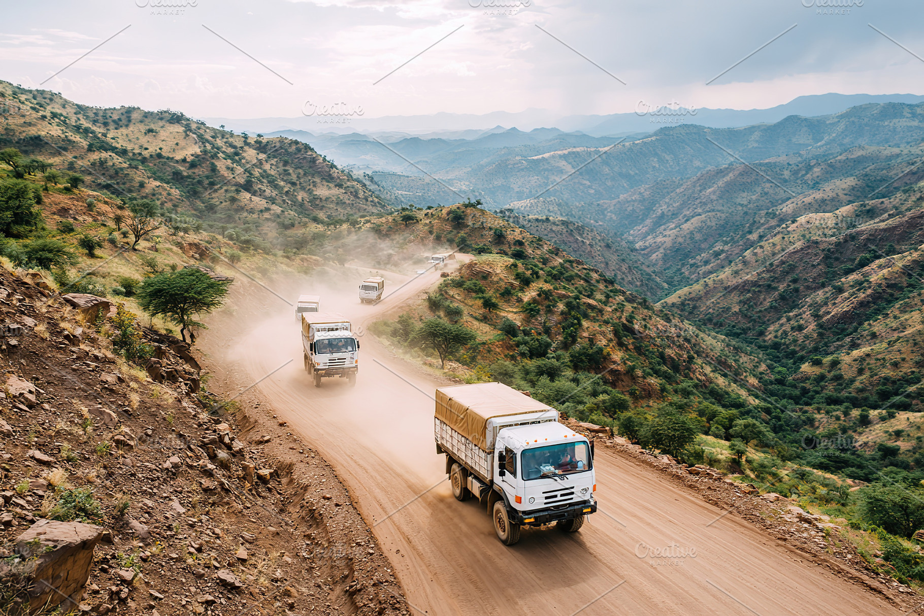 Trucks navigate a rugged mountain road during a dusty afternoon in a remote are, a Photo by AlyaAnd