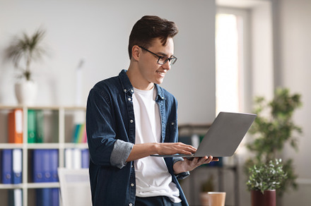 Smiling Millennial Man Using Laptop While Standing Near Desk In Modern Office