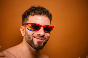 Young man smiling wearing red sunglasses with rainbow paint, a Photo by karrastock