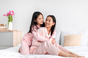 Smiling japanese teenage girl in pajama hugs young woman on bed and have fun in, a Photo by Prostock-Studio (Thumbnail 1 of 2)