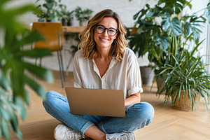 Woman sitting cross-legged on the floor using a laptop in a bright, plant-fille, a Photo by AlyaAnd