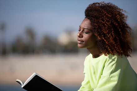 Side view of a young curly afro woman sitting on a breakwater holding a book wh