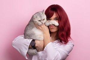 Young woman with red hair holding a playful gray cat against a p, a Person Photo by Ales Munt